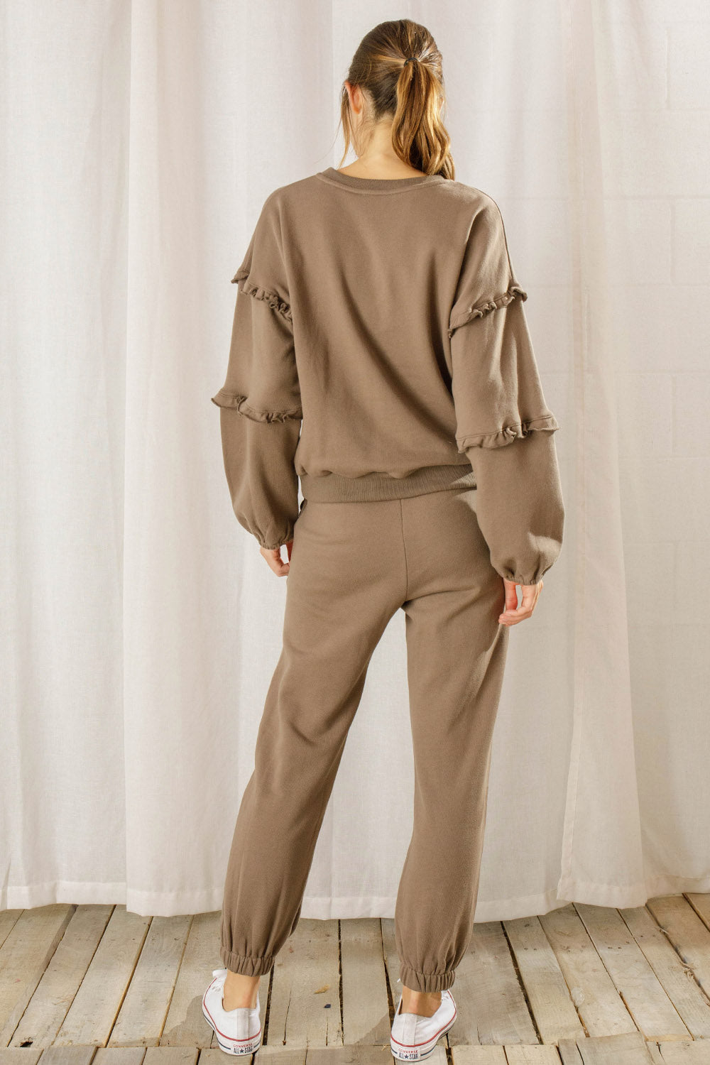 Back view of a person wearing a brown lounge set with ruffled sleeves against a white curtain background