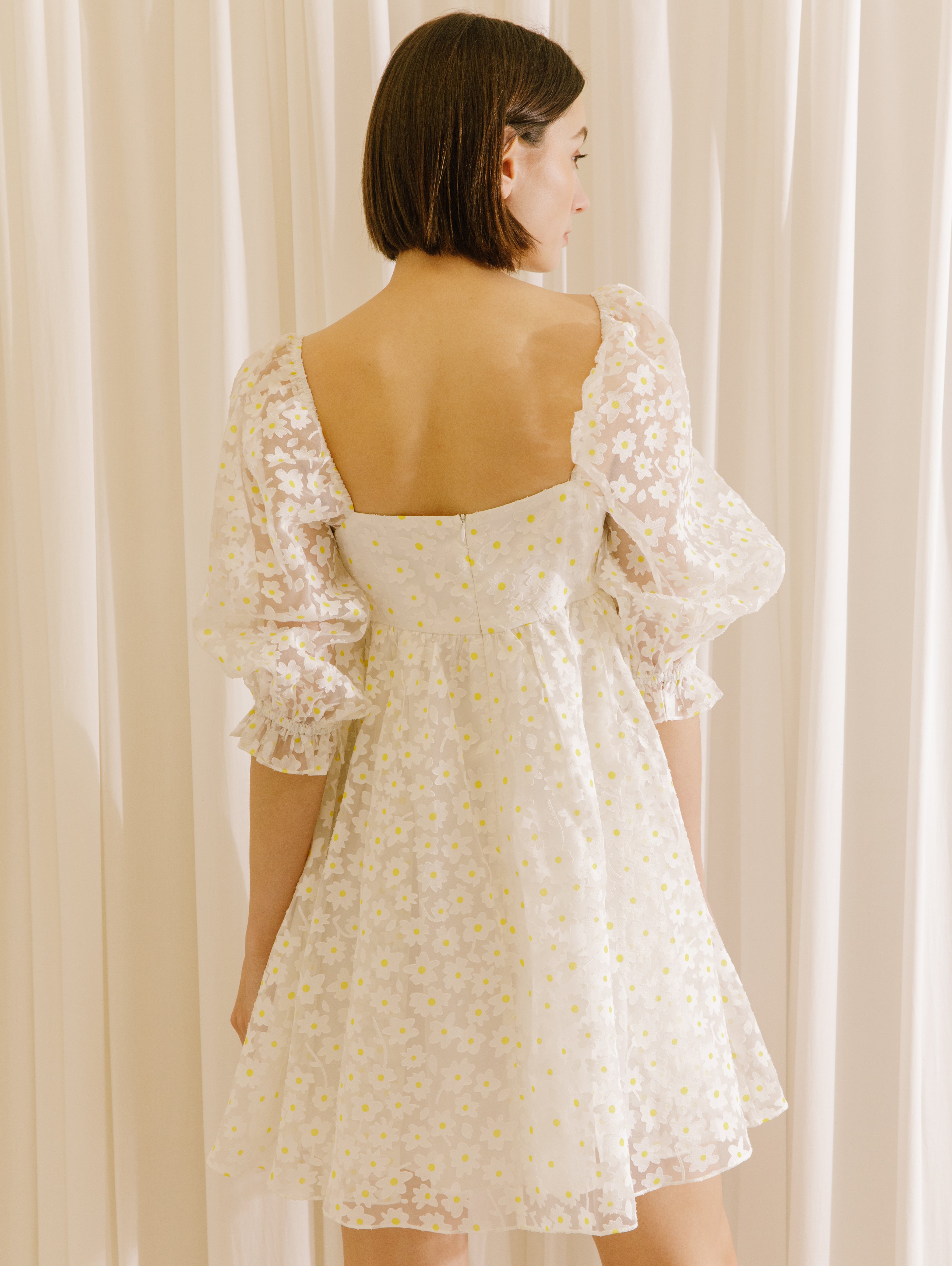Back view of a woman wearing a white dress with a sheer daisy-flocked fabric with yellow accents and a invisible zipper against a beige curtain background