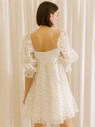 Back view of a woman wearing a white dress with a sheer daisy-flocked fabric with yellow accents and a invisible zipper against a beige curtain background