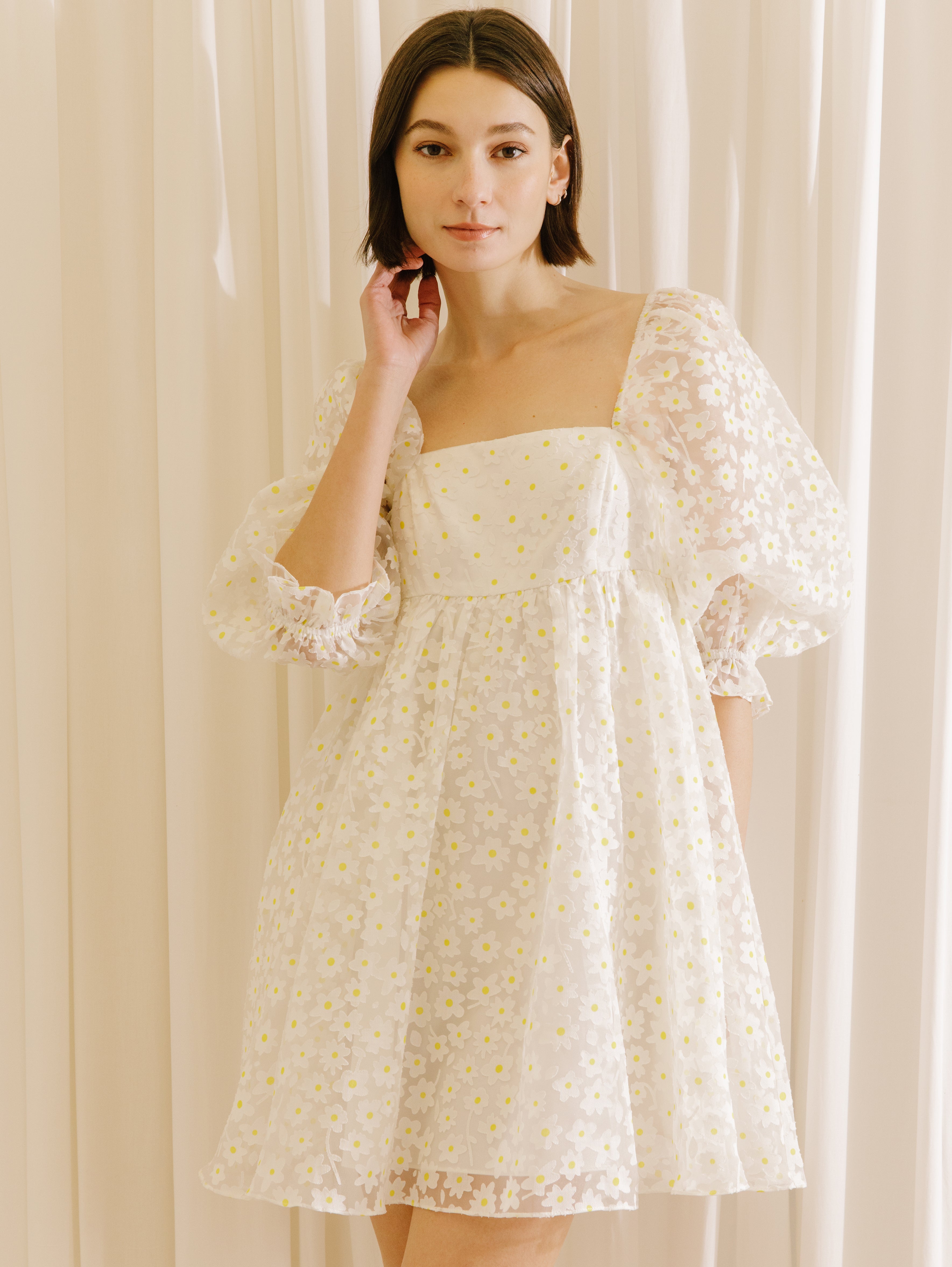 Woman wearing a white dress with a sheer daisy-flocked fabric with yellow accents against a beige curtain background