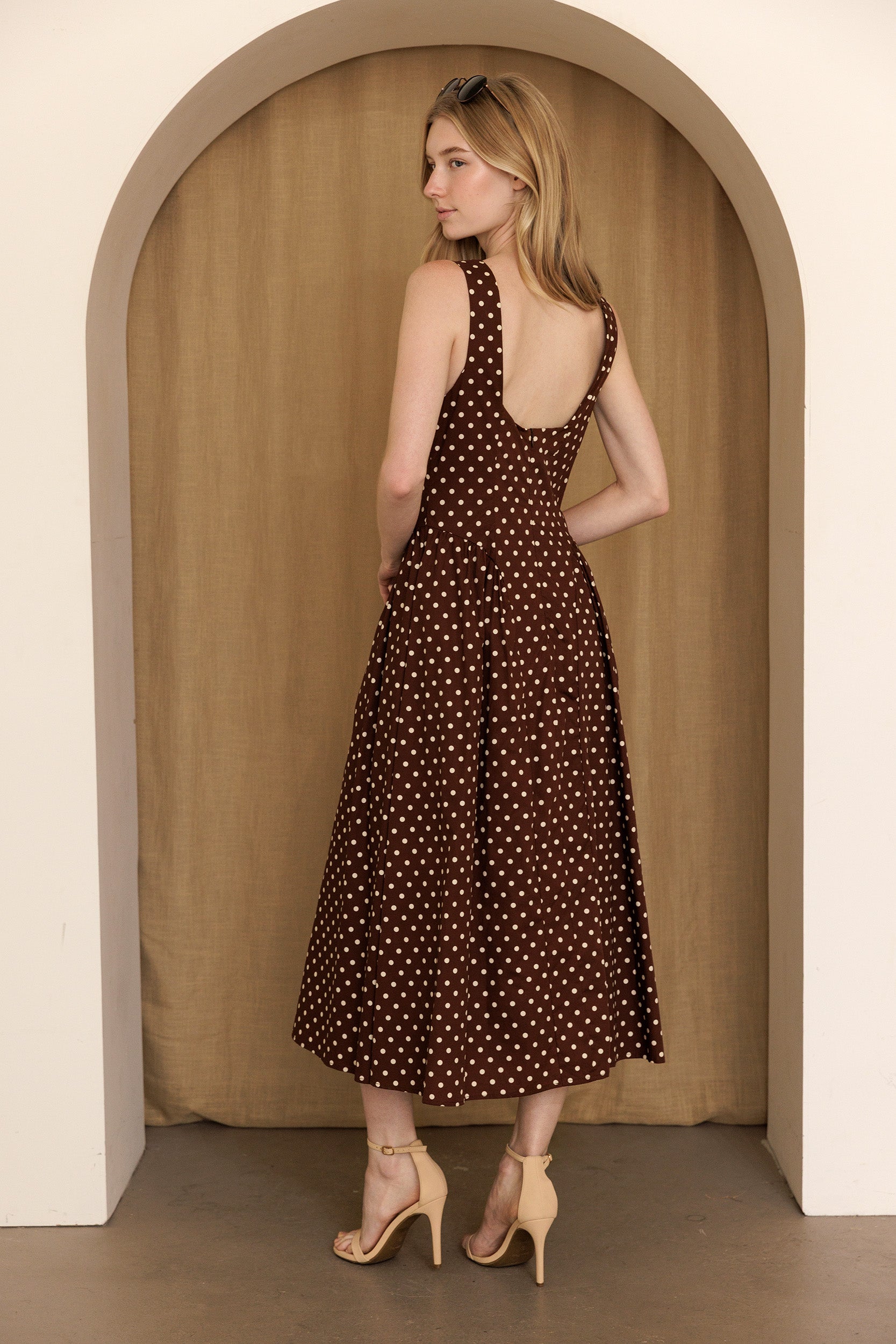 A woman viewed from behind wearing a brown polka dot midi dress standing in an archway and wearing beige high-heels.