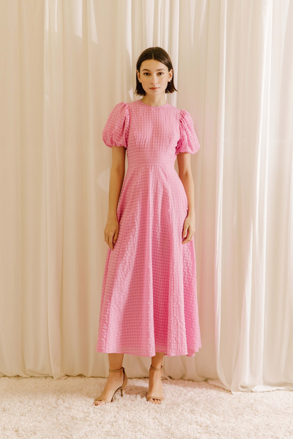 Woman wearing a pink plaid midi dress against a white curtain background