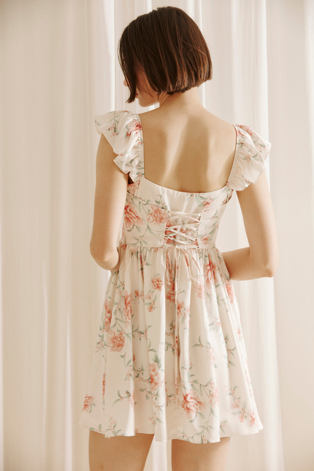 Back view of a woman wearing a cream floral mini dress with a corset crisscross back against a plain background