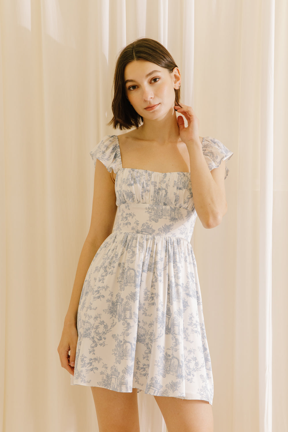 Woman wearing a white and blue floral mini dress against a beige curtain background