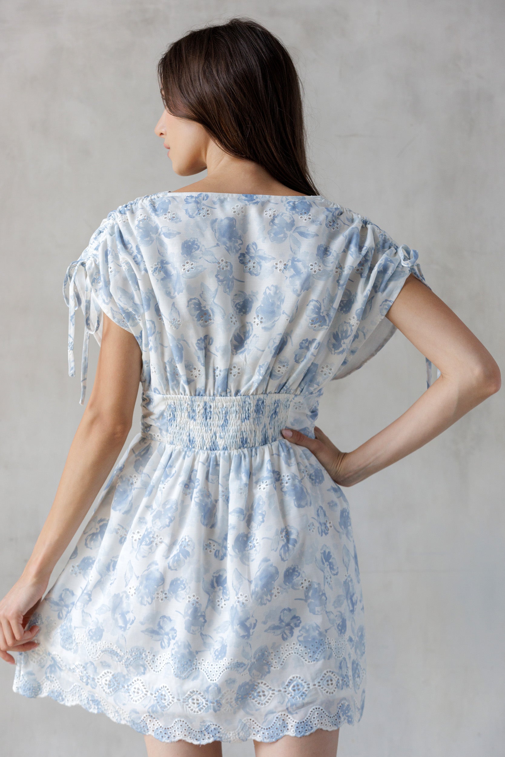 Back view of a woman wearing a blue floral dress with eyelet detailing throughout against a gray background