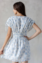 Back view of a woman wearing a blue floral dress with eyelet detailing throughout against a gray background