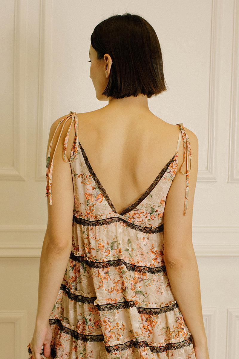 Back view of a woman wearing a peach floral dress with black lace details and self-tie straps against a neutral wall.