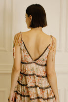 Back view of a woman wearing a peach floral dress with black lace details and self-tie straps against a neutral wall.