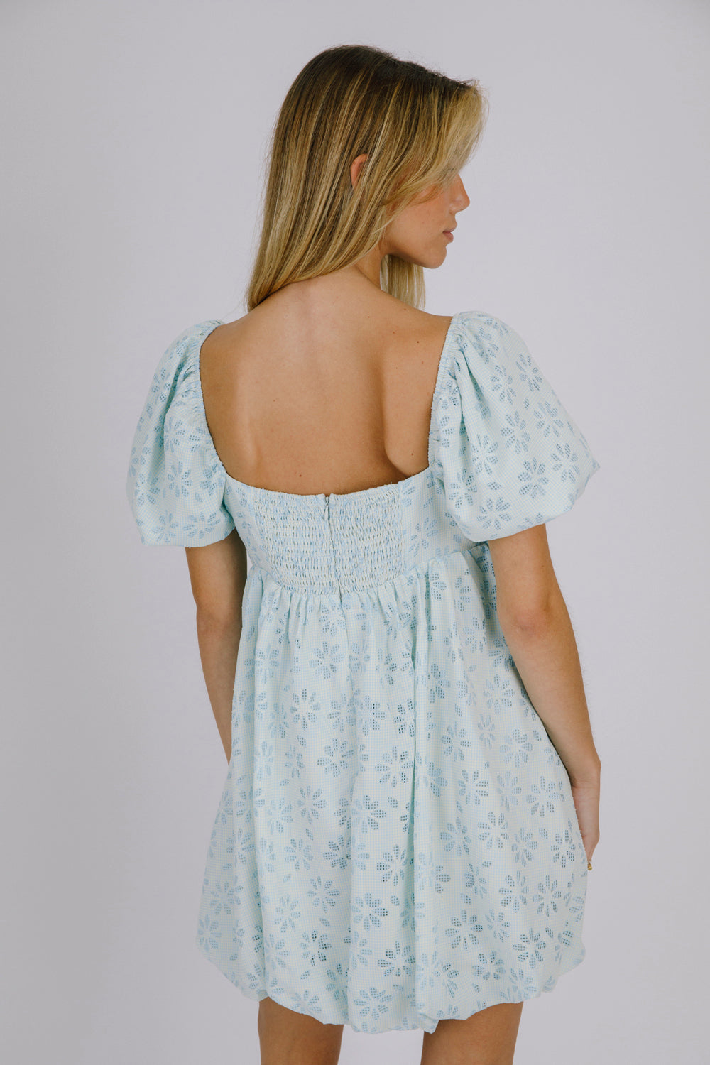 Back view of a woman wearing a blue gingham with floral embroidery mini dress against a plain background