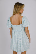 Back view of a woman wearing a blue gingham with floral embroidery mini dress against a plain background