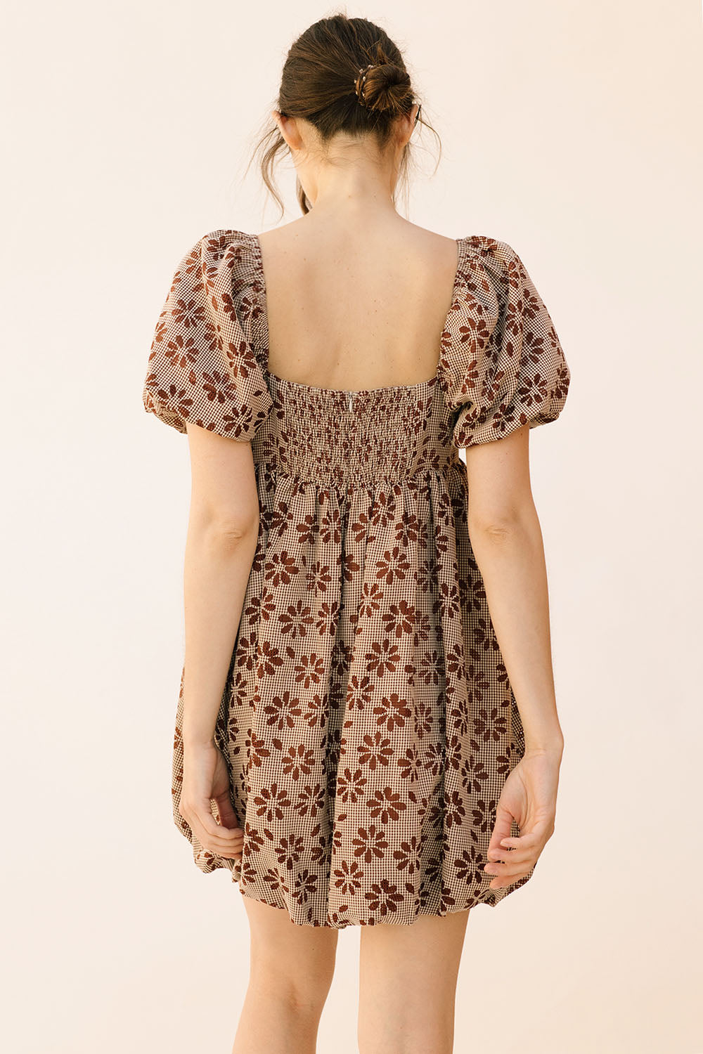 Back view of a woman wearing a brown gingham with floral embroidery mini dress against a beige background
