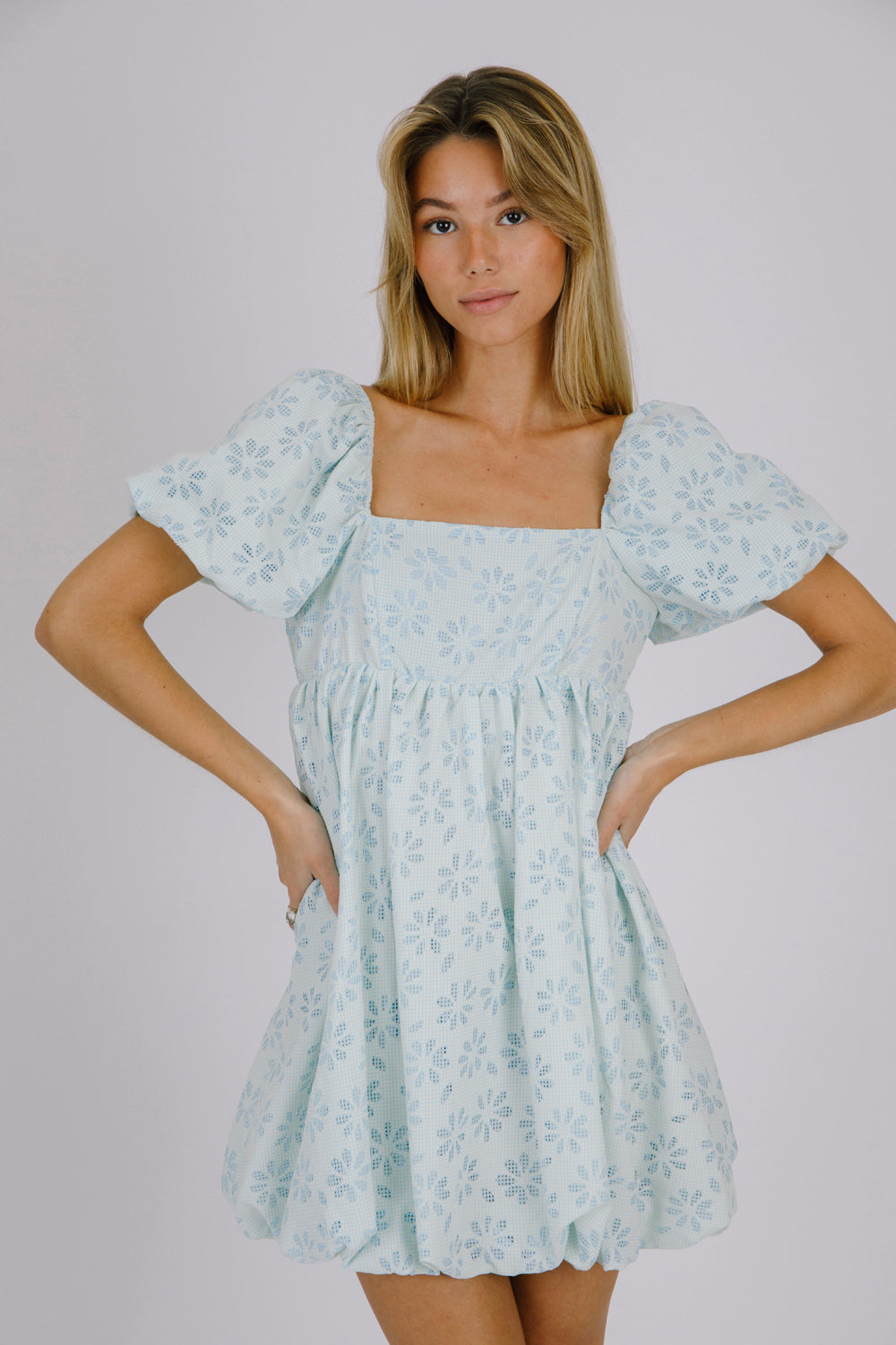 Woman wearing a blue gingham with floral embroidery mini dress against a beige background