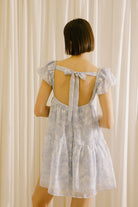 Back view of a woman wearing a light blue dress with a back self-tie bow against a beige curtain background