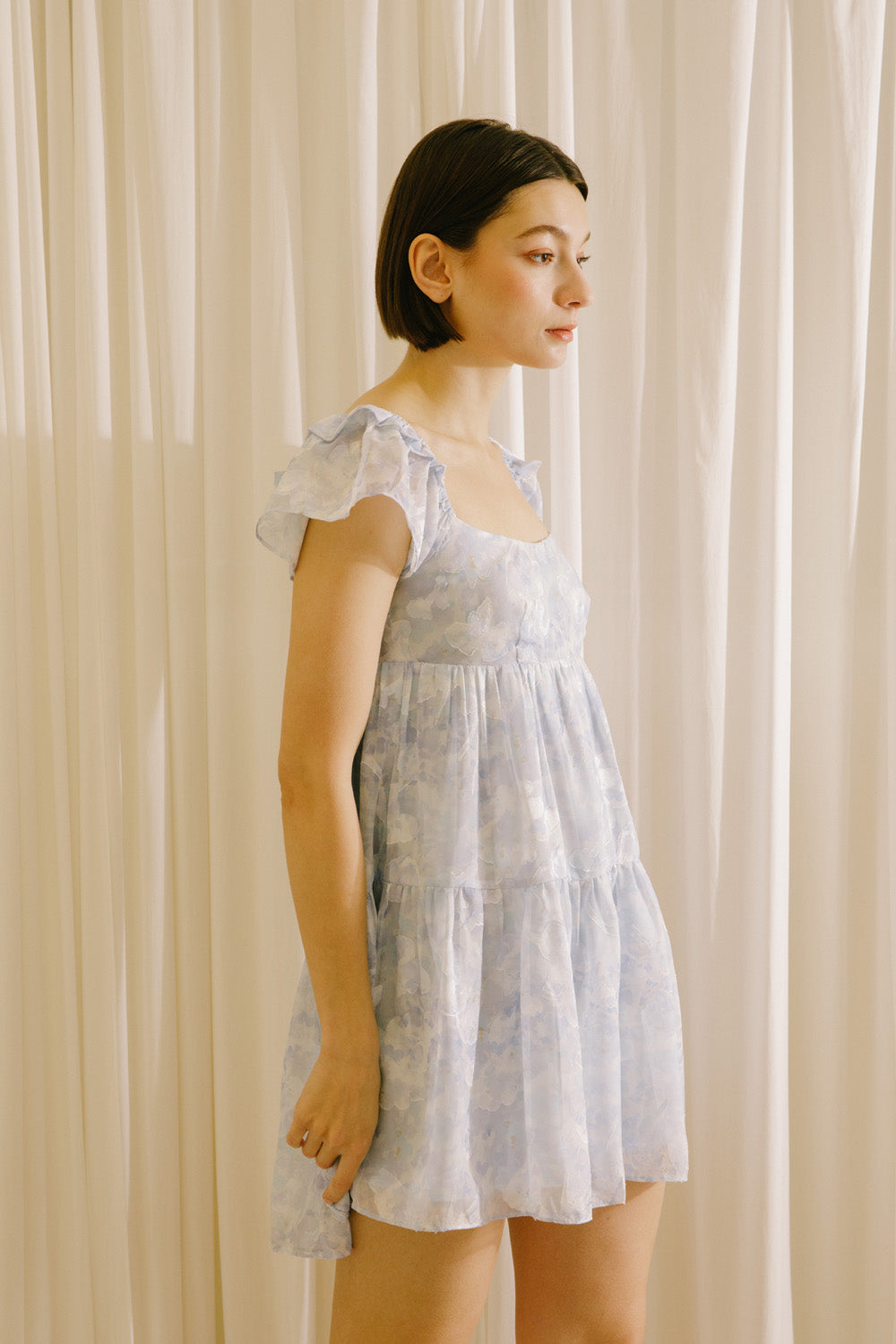 Side view of a woman wearing a light blue dress against a beige curtain background