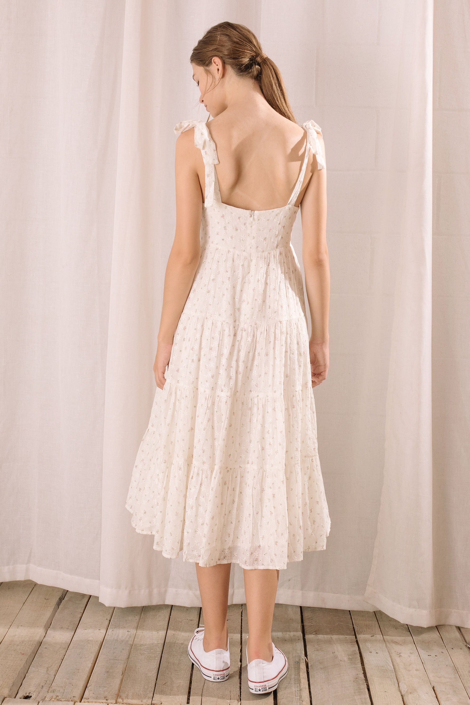 Back view of a woman wearing a white floral dress standing against a plain background