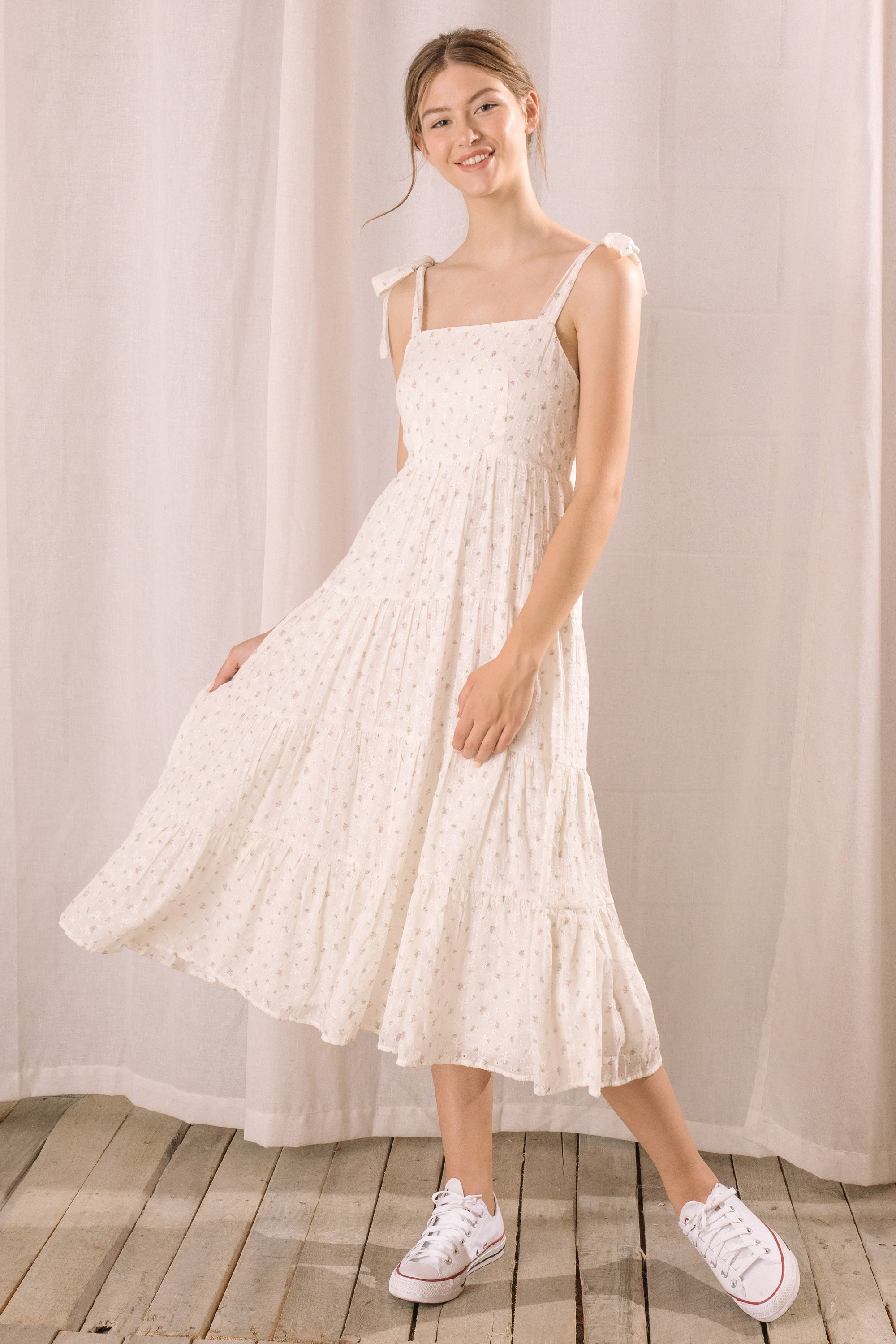 Woman wearing a white floral dress with white sneakers on a wooden floor and white curtain background