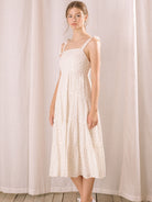 Side view of a woman wearing a white floral dress standing against a light curtain background
