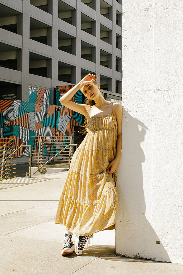 A woman standing against a white wall wearing a yellow floral ruffled maxi dress with a straight neckline, empire waist, and flowy skirt. She has her hand raised to her head.