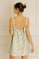 Back view of a woman wearing a green and violet dress with embroidered leave against a neutral background