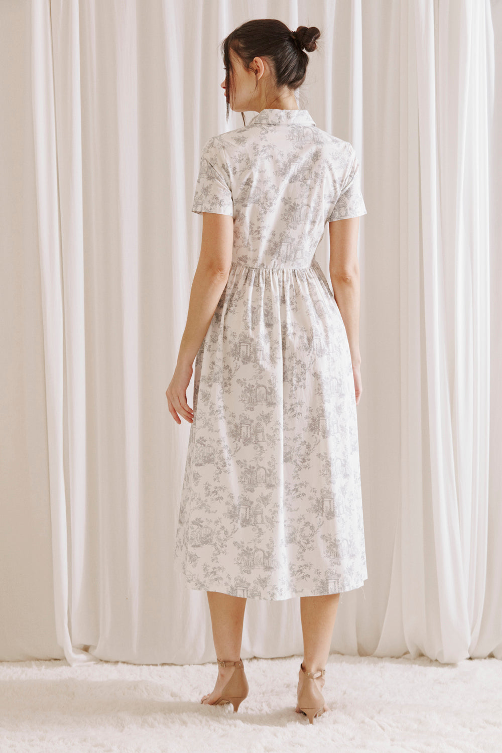 Back view of a woman wearing a white and gray toile print midi dress standing against a white curtain backdrop