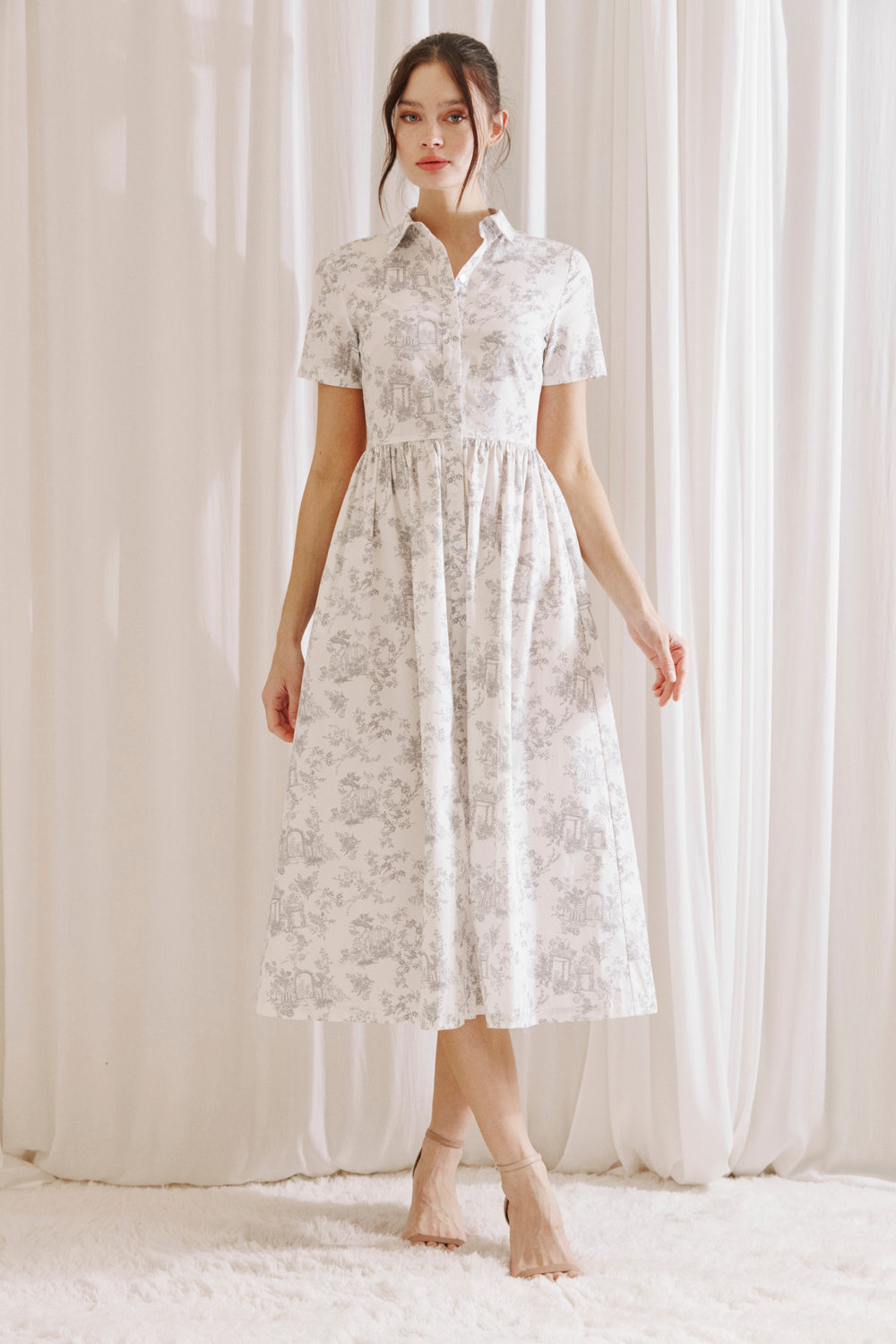Woman wearing a white and gray toile print midi dress standing against a white curtain backdrop