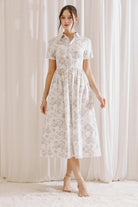 Woman wearing a white and gray toile print midi dress standing against a white curtain backdrop