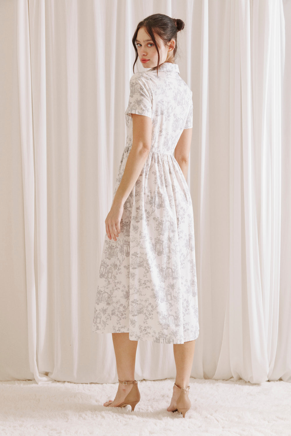 Side view of a woman wearing a white and gray toile print midi dress standing against a white curtain backdrop