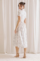 Side view of a woman wearing a white and gray toile print midi dress standing against a white curtain backdrop