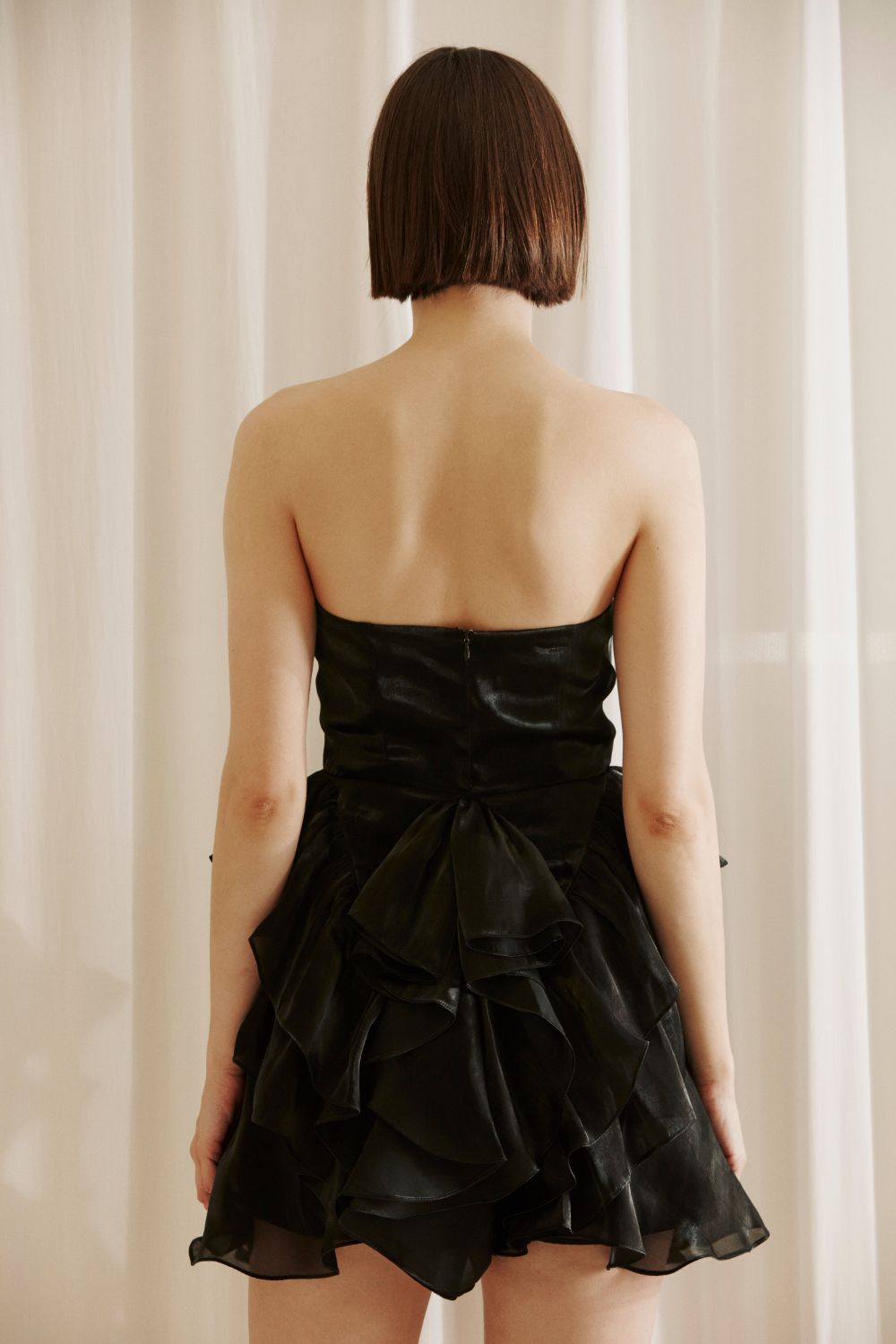 Back view of a woman wearing a black strapless dress with ruffled layers against a beige curtain background