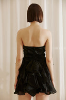 Back view of a woman wearing a black strapless dress with ruffled layers against a beige curtain background