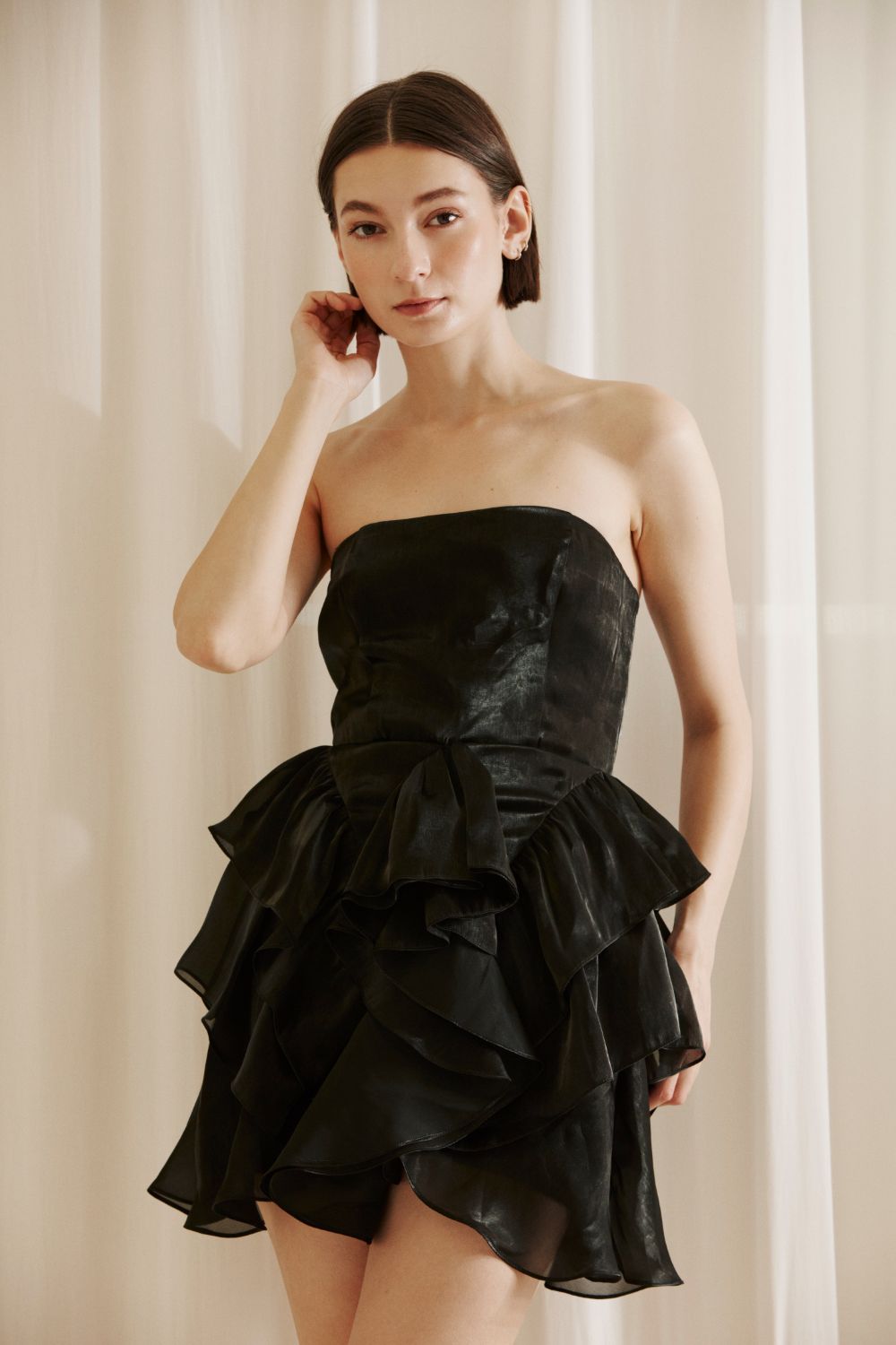 Woman wearing a black strapless dress with ruffled layers against a beige curtain background