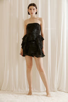 Woman wearing a black strapless mini dress with ruffled layers with heels against a beige curtain background