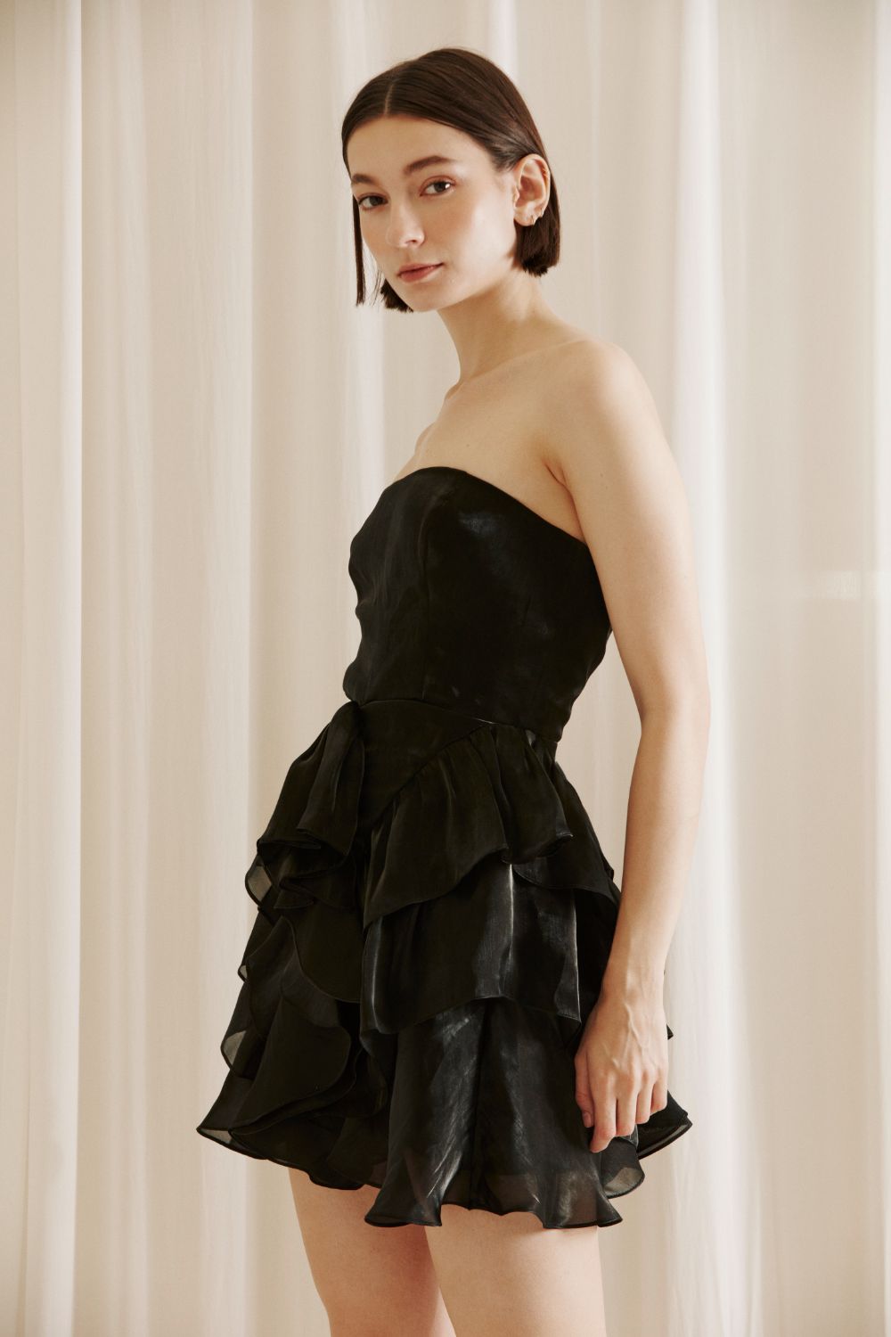 Side view of a woman wearing a black strapless dress with ruffled layers against a beige curtain background