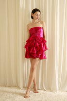 Woman wearing a bright pink strapless dress with ruffled details and beige heels against a beige wall.