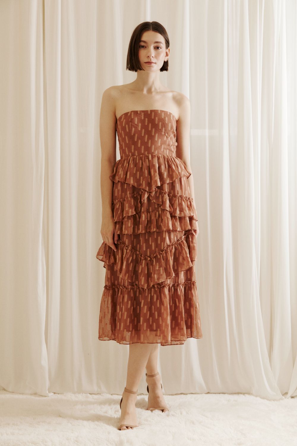 Woman wearing a strapless, tiered dress with a subtle pattern against a white curtain background