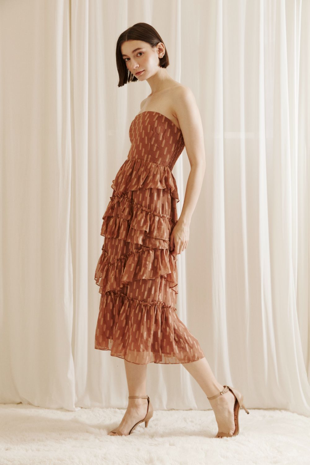 Side view of  woman wearing a strapless, tiered dress with a subtle pattern against a white curtain background