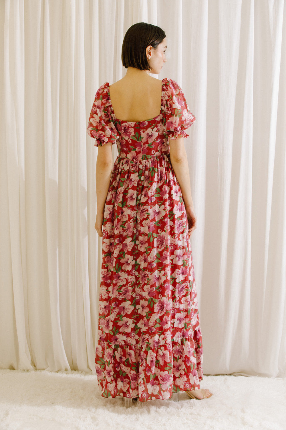 Back view of a woman wearing a berry floral dress with a slit, standing against a white curtain.