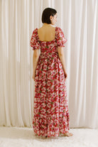 Back view of a woman wearing a berry floral dress with a slit, standing against a white curtain.