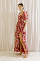 Side view of a woman wearing a berry floral dress with a slit, standing against a white curtain.