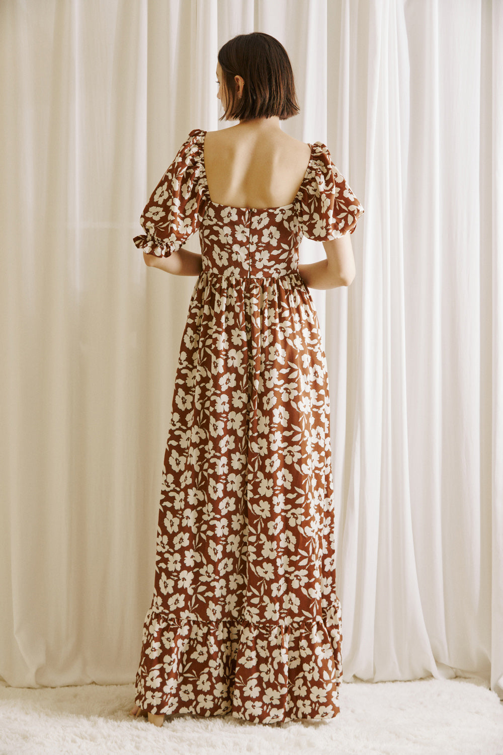 Back view of a woman wearing a brown and white floral dress standing against a white curtain backdrop