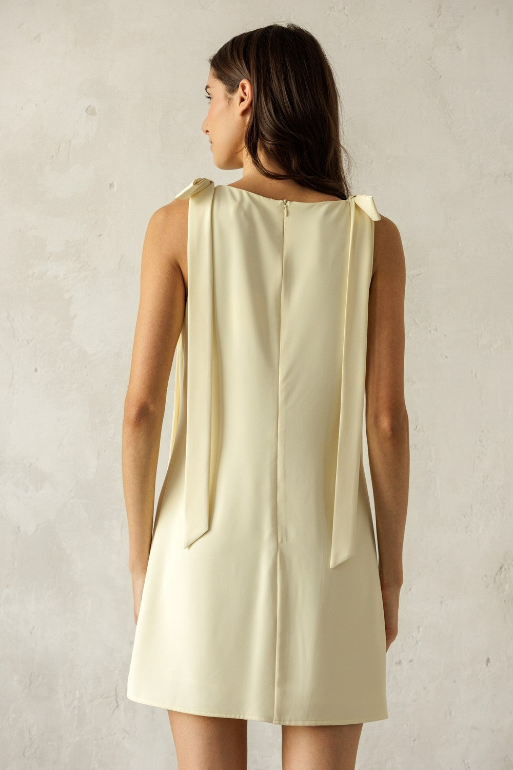 Back of a woman wearing a light yellow sleeveless dress against a plain background