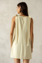 Back of a woman wearing a light yellow sleeveless dress against a plain background