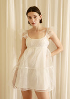 Woman wearing a white swiss dot overlay dress against a beige curtain background
