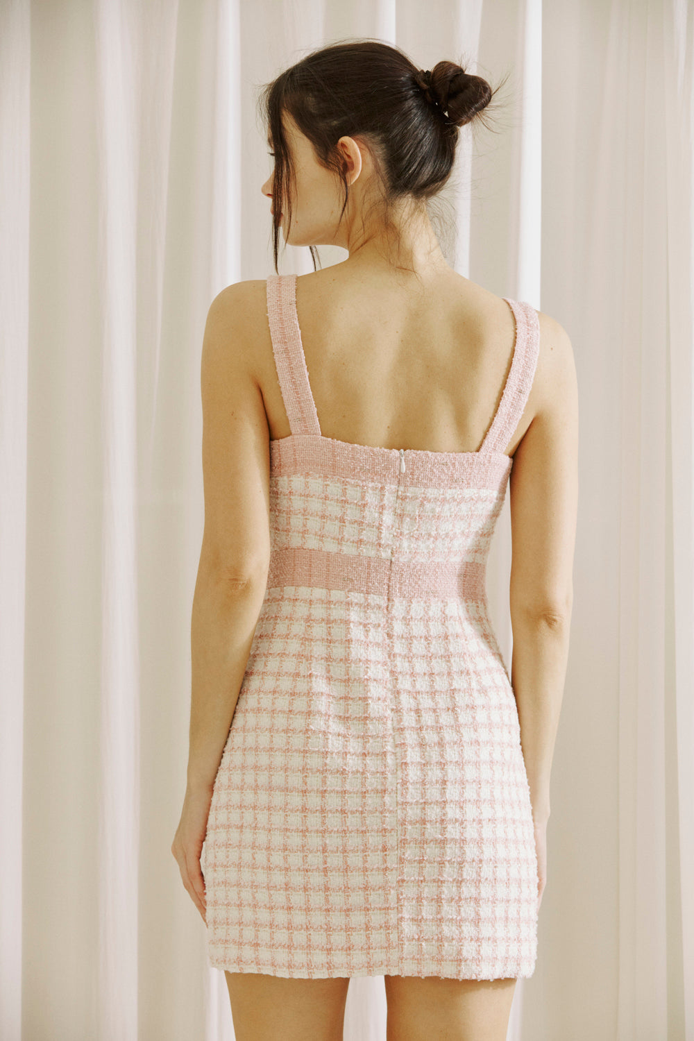 Back view of a woman wearing a pink and white checkered tweed dress against a white curtain background