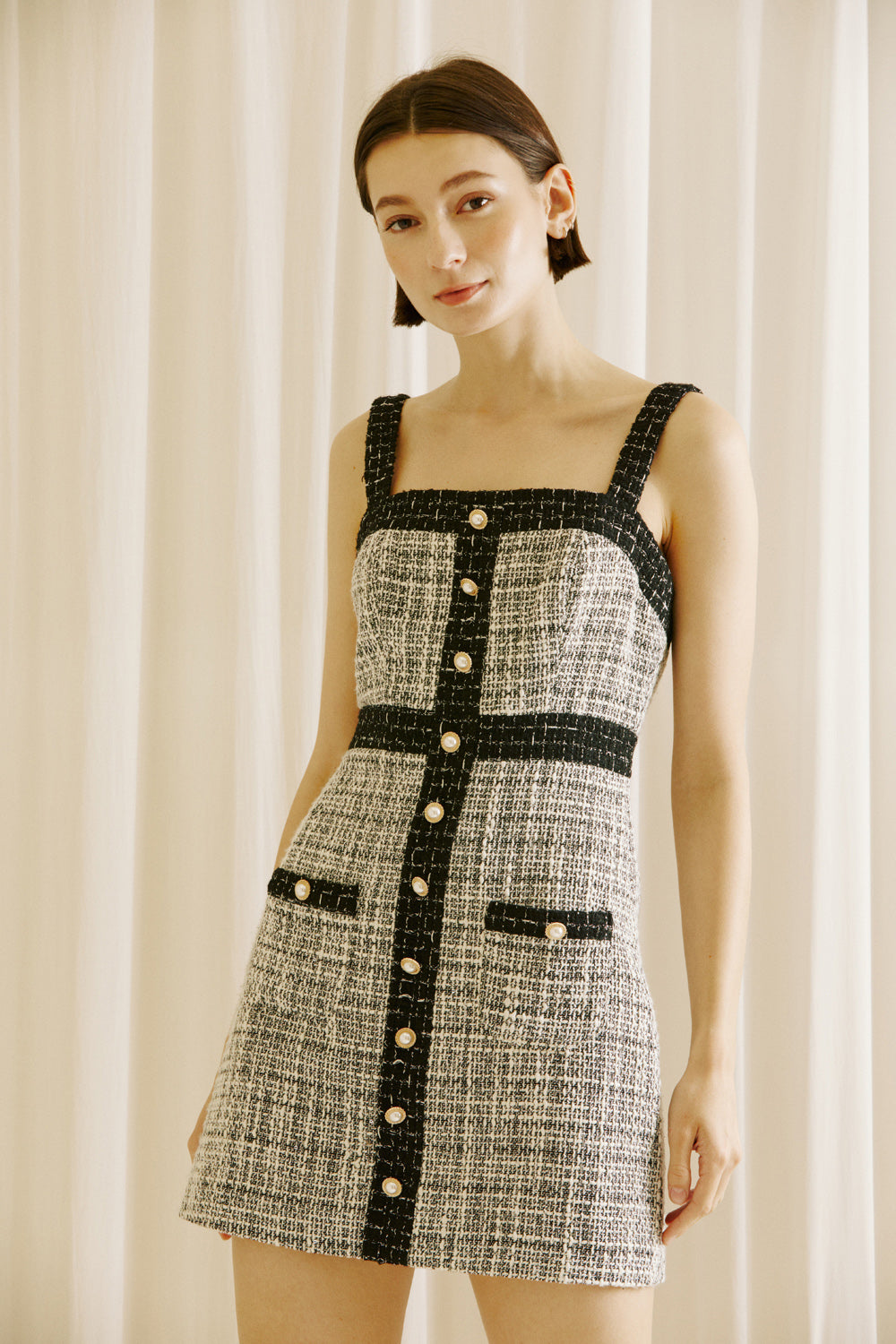 Woman wearing a black and white checkered tweed dress with pearl button details against a white curtain background