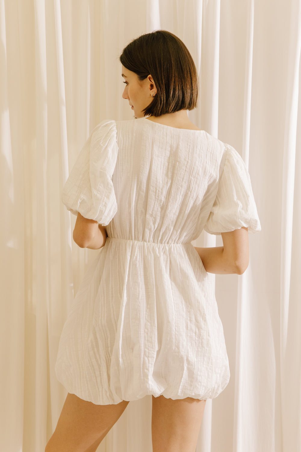 Back view of a woman wearing a white dress against a beige curtain background