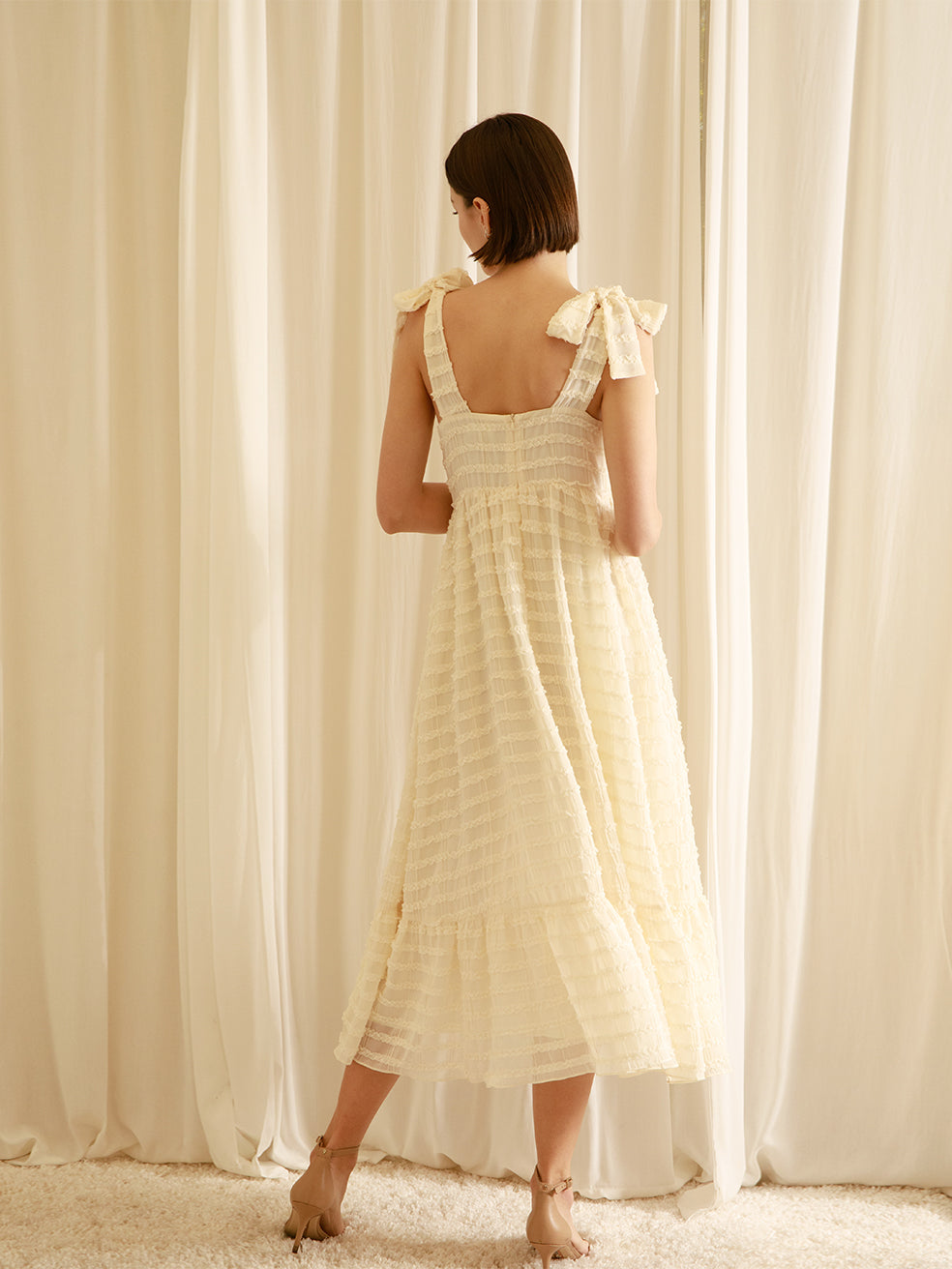 Back view of a woman wearing a cream midi dress standing in front of white curtains