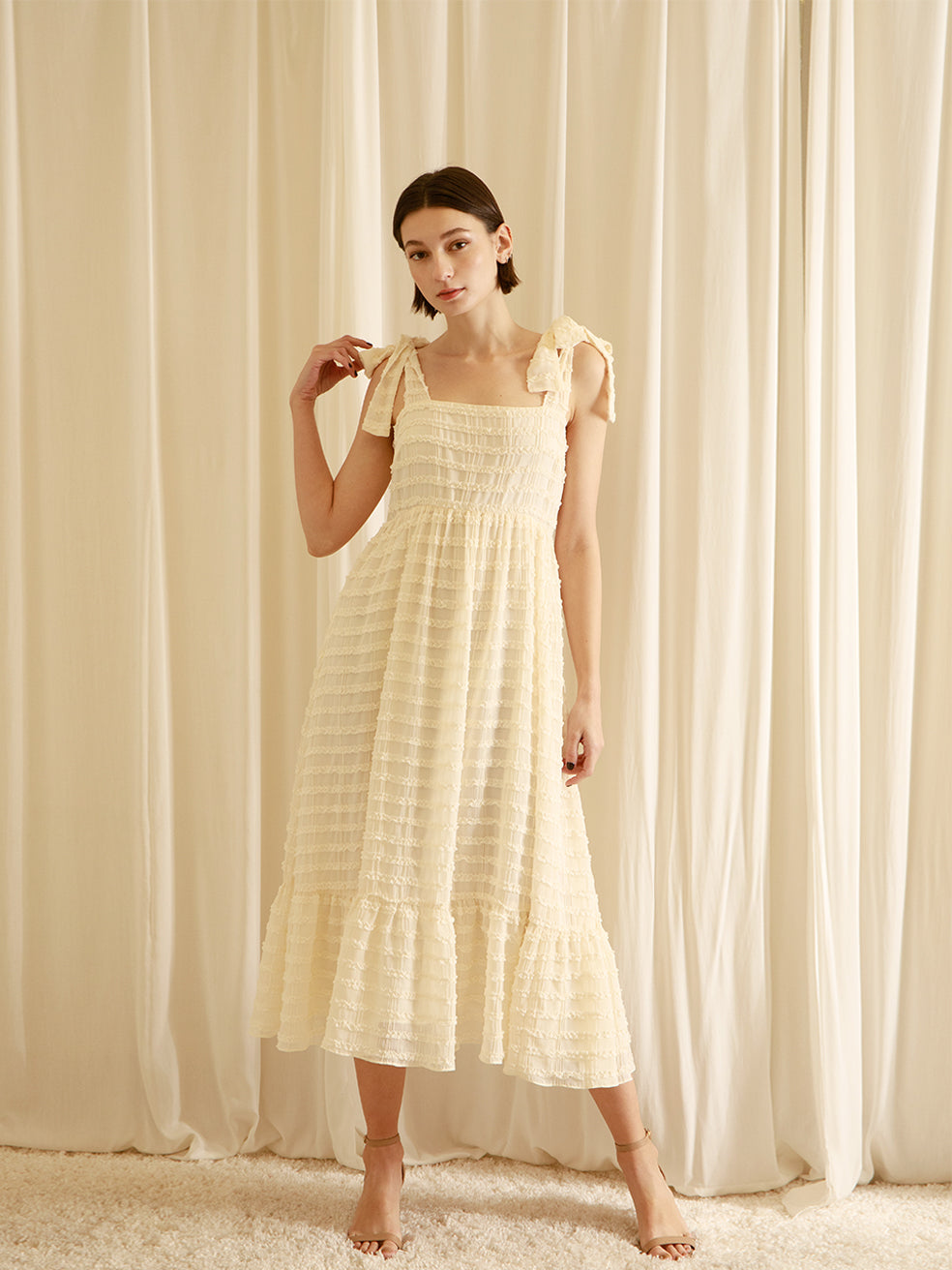 Woman wearing a cream midi dress standing in front of white curtains