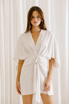 Woman wearing a white mini dress with a twisted front detail against a white curtain background