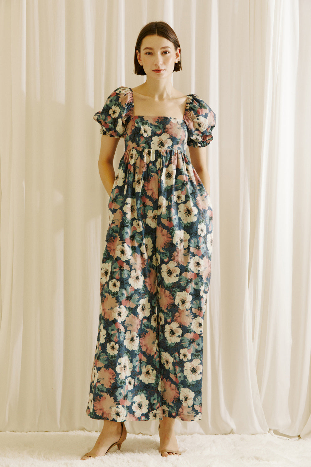 Woman wearing a flowy jumpsuit standing in front of white curtains